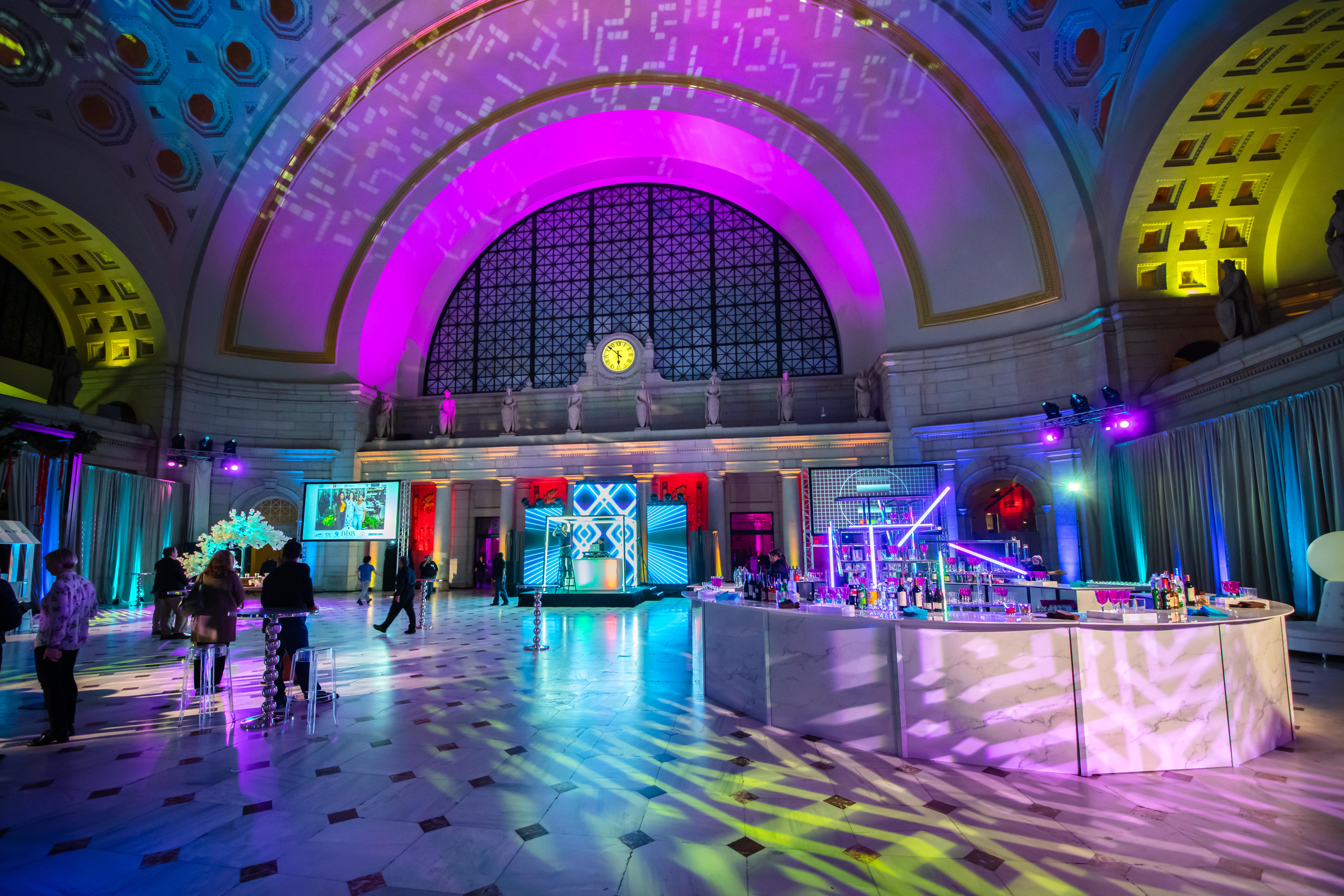 Corporate Event Cocktail Reception in Main Hall-East featuring a Stage, 360-Bar, Lounge Seating, and Neon Lights paired with Pink and Blue Up-Lighting.