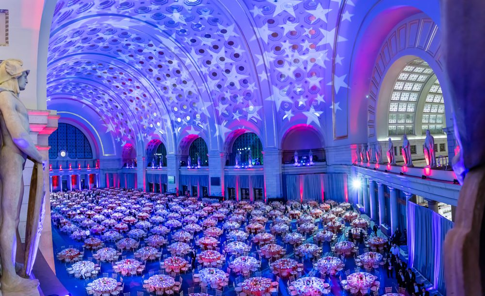 Corporate Event in Main Hall featuring a Seated Dinner; with Red and Blue Up-Lighting and White Stars Projected onto the Ceiling.