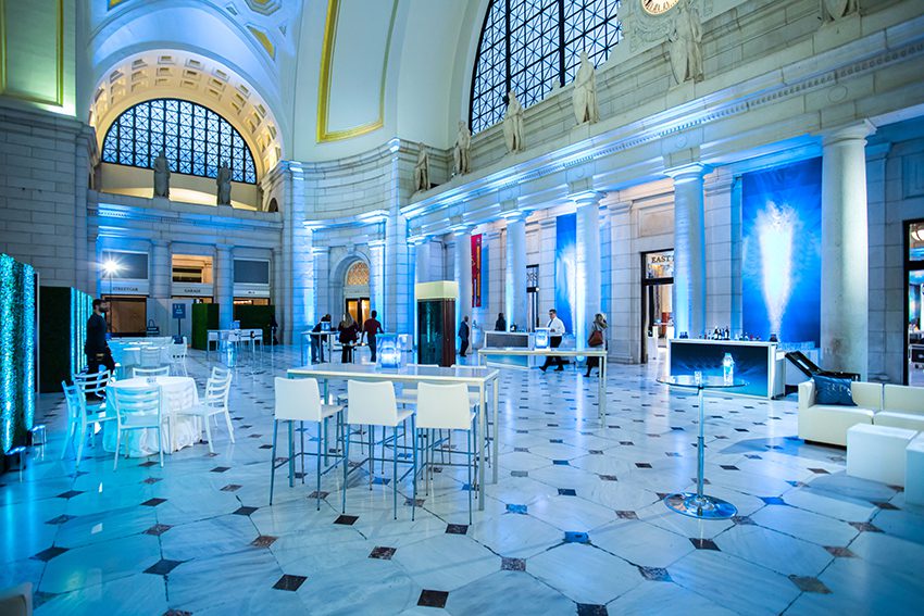 Corporate Event Cocktail Reception in Main Hall-East featuring 2 Bars, Lounge Seating, Community Tables, and Standard Seated Round Tables; with Blue Up-Lighting.