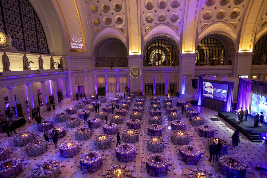 Corporate Event Cocktail Reception in Main Hall-East featuring a Stage, Buffet Stations, 360-Bar, Lounge Seating, and Neon Lights paired with Pink and Blue Up-Lighting.